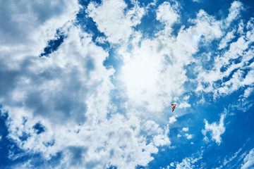 A large kite floats high in the sky among the clouds against the blue sky and the bright summer sun and it is controlled by two white ropes going to the ground