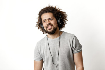 Wall Mural - A curly-headed handsome man wearing a gray T-shirt and ripped jeans is standing and listening to music in the headphones with a soft smile over the white background.