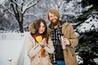 © Elizaveta - Young loving couple caucasian man with blond long hair and beard, beautiful woman have fun drinking a hot drink from thermos, eating green apple in snow park near white fence and coniferous tree.