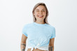 © Cookie Studio - Waist-up shot of carefree emotive young woman with braces and tattoos, smiling joyfully and winking flirty at camera, sticking out tongue, being in great mood while standing over gray wall