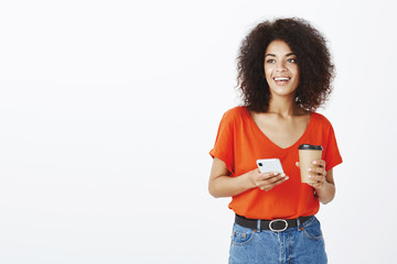 Carefree friendly-looking positive woman with dark skin and curly hair, holding cup of coffee and smarpthone, smiling and gazing aside with dreamy look, thinking or planning trip with friends