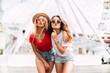 © chichkaua - Best friend having fun near Ferris wheel, going crazy together, wearing hats and mirrored sunglasses, amazing view on the city, bright colors evening sunlight
