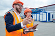 © Seventyfour - Portrait of production foreman wearing hardhat and holding clipboard talking to female factory employee outdoors while pointing sideways, copy space