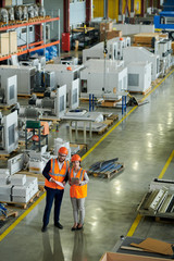 Wall Mural - High angle full length portrait of bearded businessman wearing hardhat inspecting production workshop accompanied by female factory employee, copy space