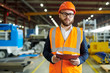 © Seventyfour - Waist up portrait of modern bearded businessman wearing hardhat and reflective vest looking at camera while posing in production workshop, holding clipboard, copy space