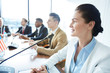 © pressmaster - Young elegant female delegate sitting by table with microphone in front and speaking at conference