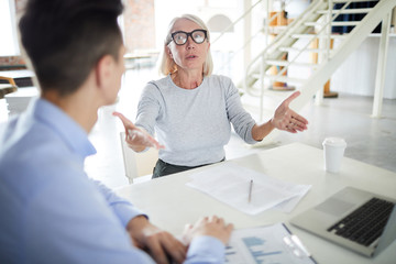 Mature female leader wearing eyeglasses sitting at desk and gesturing during a business meeting at office