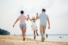 Back View Of Modern Couple In Casualwear Lifting Their Little Daughter By Hands While Slightly Lifting Her Over Sand During Chill On The Beach