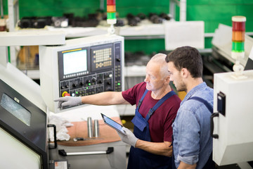 Mature foreman controlling the process of machine on his tablet pc and showing how its work to the worker