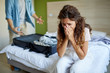© pressmaster - Young woman sitting on bed and crying while her angry husband getting ready with suitcase