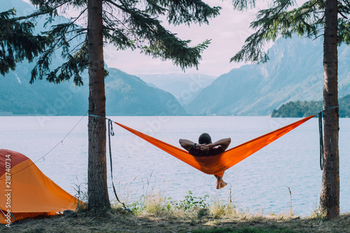 Photographie Back view of man silhouette relaxing on orange hammock between two trees pine enjoying the view at the lake in summer norwegian cloudy morning
