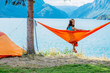 © Iryna - Young barefoot woman turn around while relaxing on orange hammock between two trees pine enjoying the view at the lake in summer norwegian cloudy morning. Rest on camping concept.