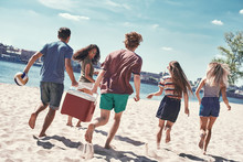 Friends On The Beach. Rear View Of Cheerful Young People Walking By The Beach To The Sea While Two Men Carrying Plastic Cooler
