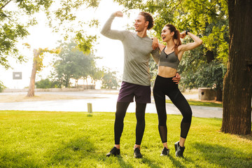 Canvas Print - Photo of joyful sportive people man and woman 20s in sportswear, training together and showing biceps in green park during sunny summer day