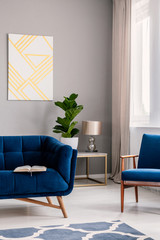 Real photo of light grey living room interior with fresh plant, window with drapes, geometric painting and open book placed on dark blue sofa