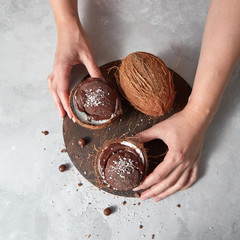 Wall Mural - A girl hold two halfs of coconut with chocolate sorbet with coconut chips and chocolate balls on a wooden board on a gray concrete. Top view.