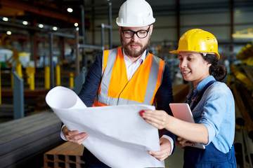 Wall Mural - Portrait of modern bearded engineer wearing hardhat holding blueprints  while discussing production with female worker in factory workshop, copy space