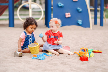 Innocent look. Two cute Caucasian and hispanic latin babies children sitting in sandbox playing with plastic colorful toys. Little girls friends having fun together on playground.