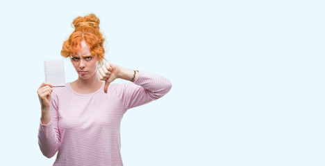 Poster - Young redhead woman holding blank notebook with angry face, negative sign showing dislike with thumbs down, rejection concept