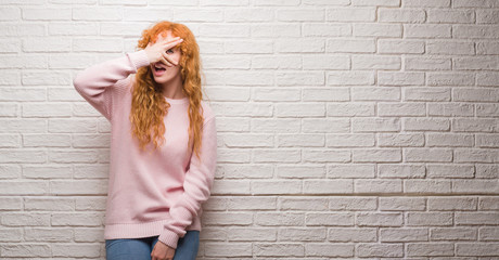 Wall Mural - Young redhead woman standing over brick wall peeking in shock covering face and eyes with hand, looking through fingers with embarrassed expression.