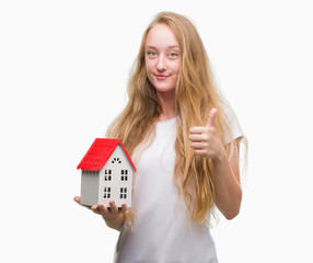 Sticker - Blonde teenager woman holding family house happy with big smile doing ok sign, thumb up with fingers, excellent sign