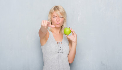 Adult caucasian woman over grunge grey wall eating green apple pointing with finger to the camera and to you, hand sign, positive and confident gesture from the front