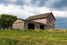 Abandoned Farm With Old Painted Red Barns Standing Empty On A Hill With Dark Clouds. Gives The Feeling Of Trouble, A Storm Brewing For Farmers. Concepts Of Family Farm, Trade Wars, Tariffs