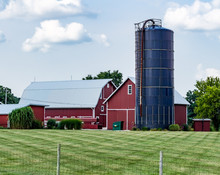 Modern, Bright Red Barn, Farm Buildings And Blue Silo, With Blue Sky And Puffy Clouds. Working Family Farm With Beautiful Barns And Lawn