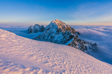 Wall Mural - Sunset view of a winter alpine like landscape. Vibrant blue sky, purple and pink color on low clouds and snow. Sharp rocky summit and peak of Pysny and Kezmarsky stit in High Tatras, Slovakia.