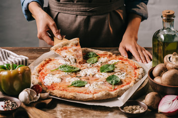 Wall Mural - partial view of woman in apron taking piece of pizza on cutting board on wooden tabletop