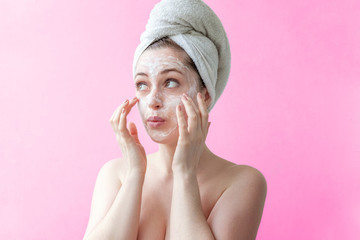 Beauty portrait of a smiling brunette woman in a towel on the head applying white nourishing mask or creme on face on pink background isolated. Skincare cleansing spa relax concept