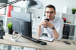 © Ivan Traimak - A young man in glasses stands near a table in the office holding a glass of coffee.