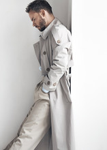 Fashion portrait of man with dark beard and hair, weared in light trench coat and beige pants.