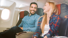 On A Commercial Plane Flight Handsome Hispanic Man Tells Funny Story To His Beautiful Blonde Girlfriend. Both Laugh. They Travel In New Airplane, With Sun Shining Through The Window.