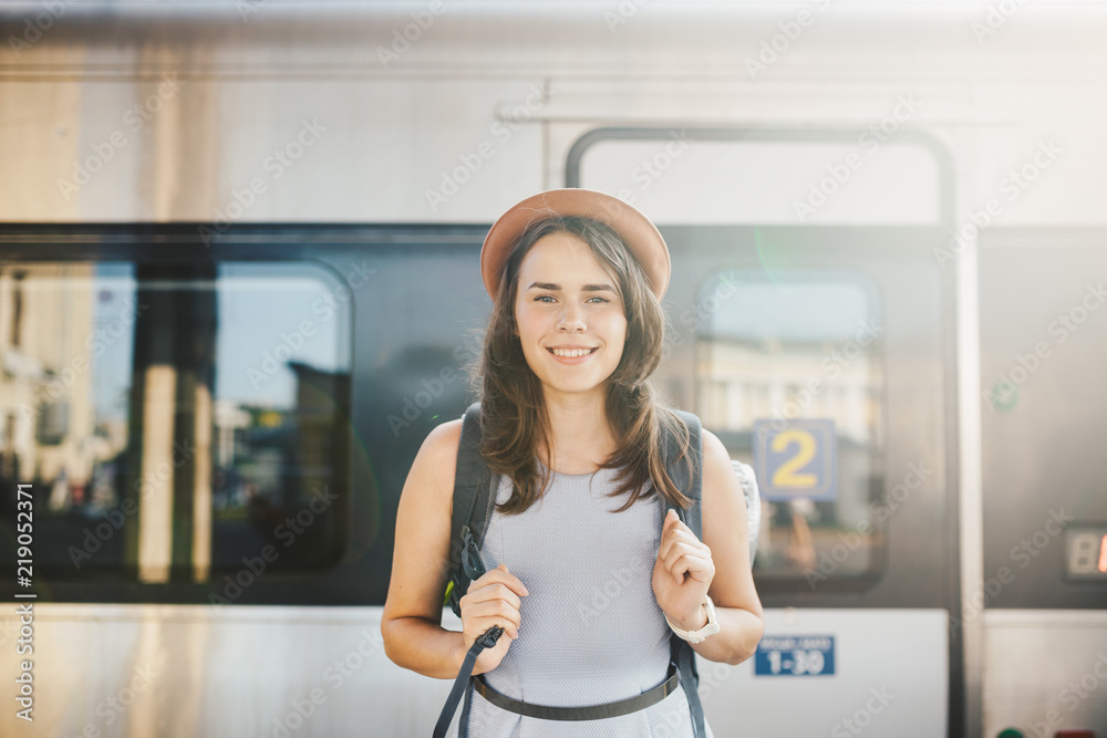 theme railway and travel. Portrait young caucasian woman with toothy smile standing at train station train background with backpack and equipment for tourism isolated mat in dress and hat in summer