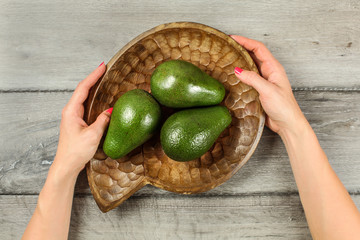 Tabletop view on gray wood desk, woman hands holding wooden carved bowl with three whole green avocados.