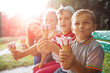 Group of four happy children eating ice cream together outdoor. Photo of happy blond girls with two handsome boys sitting on the bench and smiling at camera. Sun glare effect