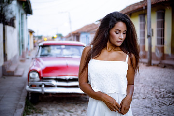 Beautiful young lady in white dress standing thoughtful in front of an old classic car in the old streets of Trinidad in Cuba.