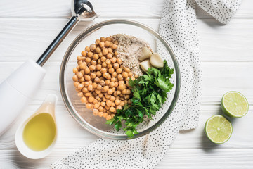 Wall Mural - Flat lay with chickpeas, parsley and spices ingredients for hummus in bowl, blender and lime pieces on wooden surface