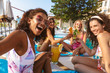 © Drobot Dean - Group of women friends sitting outdoors in pool dressed in swimwears take a selfie by camera.