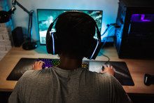 Back View Of Teenage Gamer Boy Playing Video Games Online On Computer In Dark Room, Wearing Headphones With Microphone And Using Backlit Colorful Keyboard