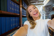 © Suzi Media  - Excited young girl standing in traditional old library at bookshelves, smiling and laughing student making selfie on phone camera, having fun. Higher education and Student life concept.