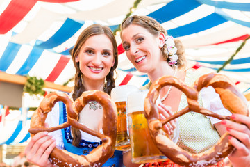 Two friends wearing dirndl are standing in a beer tent at Dult or Oktoberfest with giant pretzels