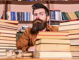 Leinwandbilder - Man on strict face sit between piles of books, while studying in library, bookshelves on background. Teacher or student with beard sit at table with glasses, defocused. Bookworm concept.