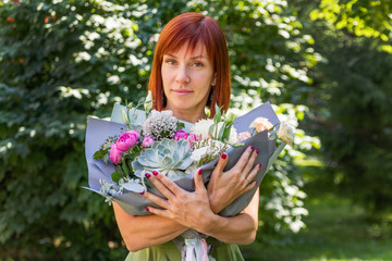 Portrait of a charming red-haired girl who stands in the park on a warm spring day with a large bouquet of scraps of flowers