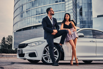 Well-dressed attractive couple leaning on a luxury car outdoors against the skyscraper.