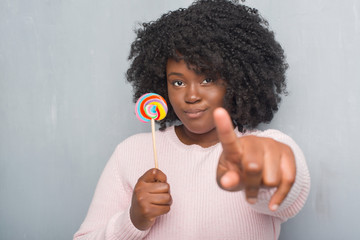 Wall Mural - Young african american woman over grey grunge wall eating lollipop candy pointing with finger to the camera and to you, hand sign, positive and confident gesture from the front