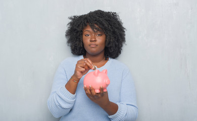 Sticker - Young african american woman over grey grunge wall holding piggy bank with a confident expression on smart face thinking serious