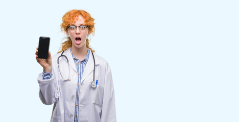 Wall Mural - Young redhead doctor woman showing smartphone scared in shock with a surprise face, afraid and excited with fear expression