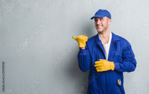 Fotomural Young caucasian man over grey grunge wall wearing mechanic uniform pointing and
